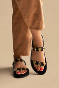 Black suede flat sandals with two straps decorated with gold and blue stone charms, front view on a model in tan trousers