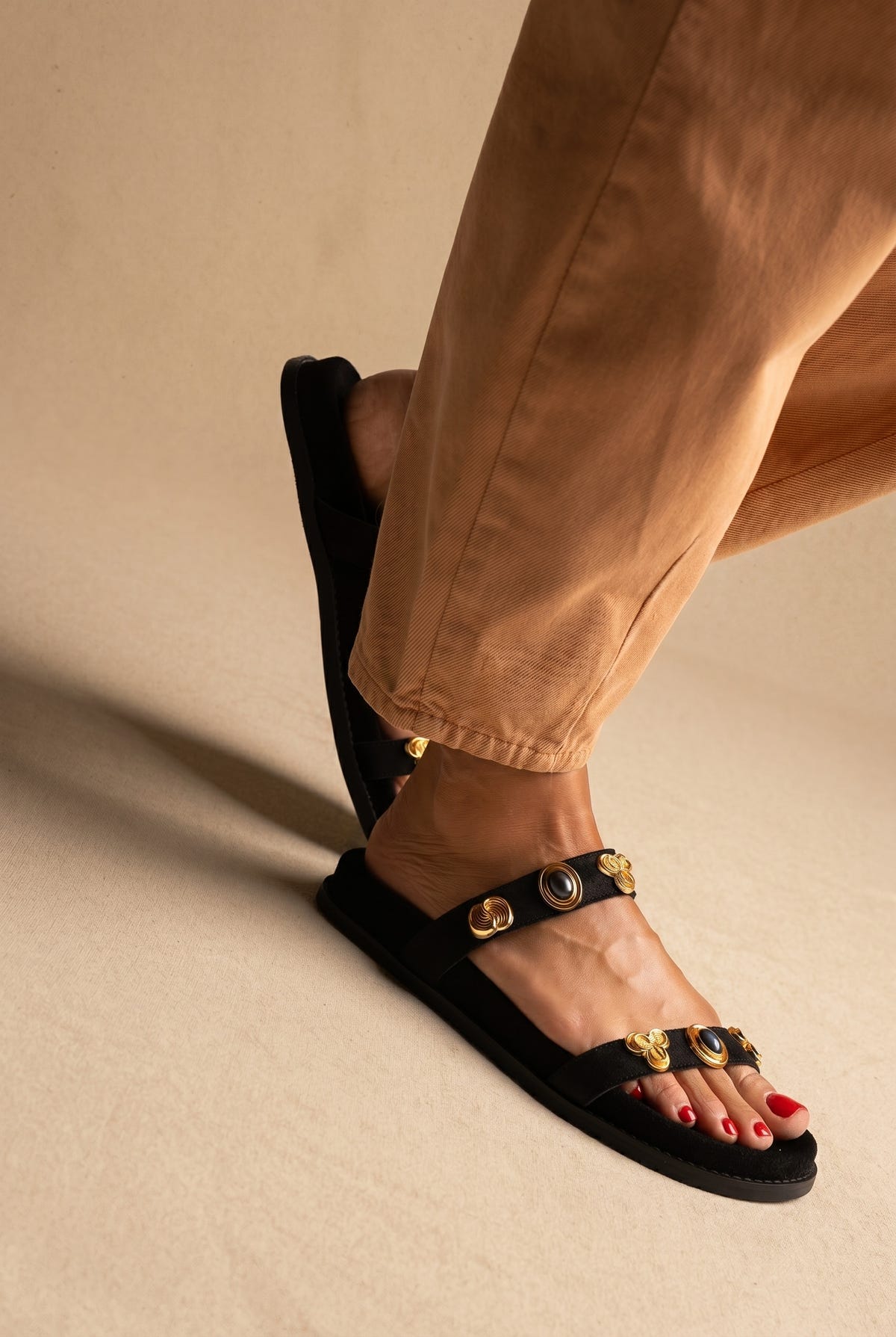 Black suede flat sandals with two jewelled gold-tone strap details, three-quarter view on a model in tan trousers