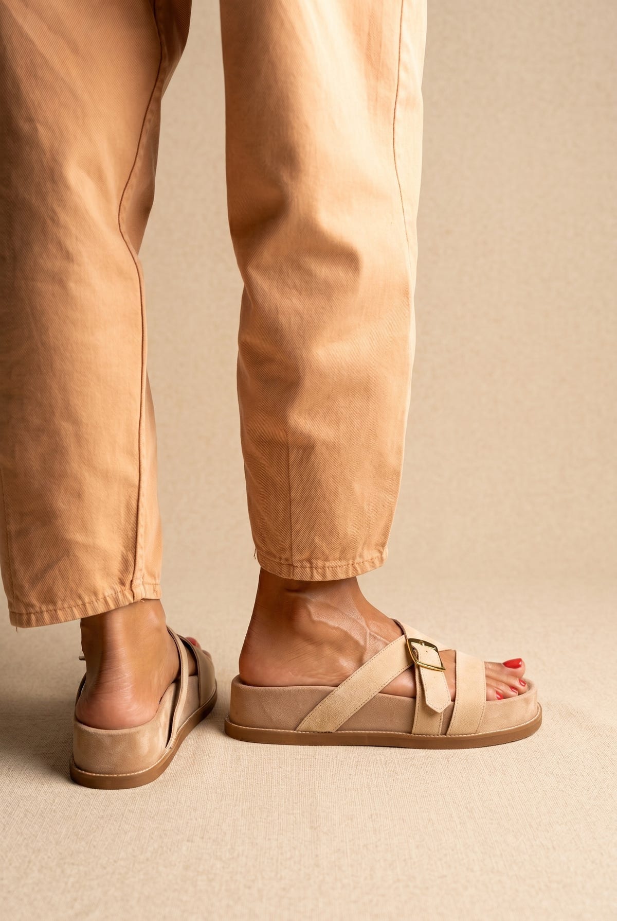 Back view of beige suede crossover-strap slide sandals with a gold buckle and chunky sole, worn on a model