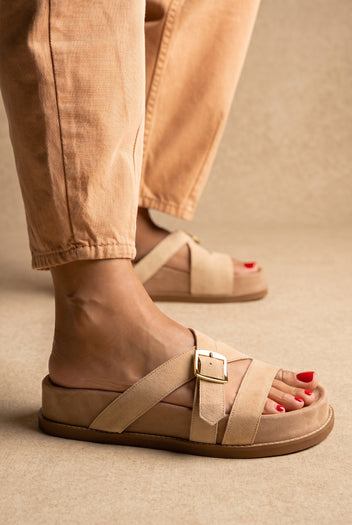 Beige suede crossover-strap slide sandals with a gold buckle and chunky sole, side view on a model in tan trousers