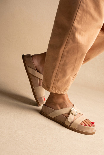Beige suede slide sandals with crossing straps and a gold buckle, worn on a model with one leg raised