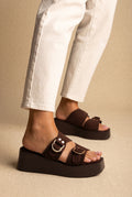 Dark brown suede platform slides with two buckled straps and gold rings, worn on a model with white jeans