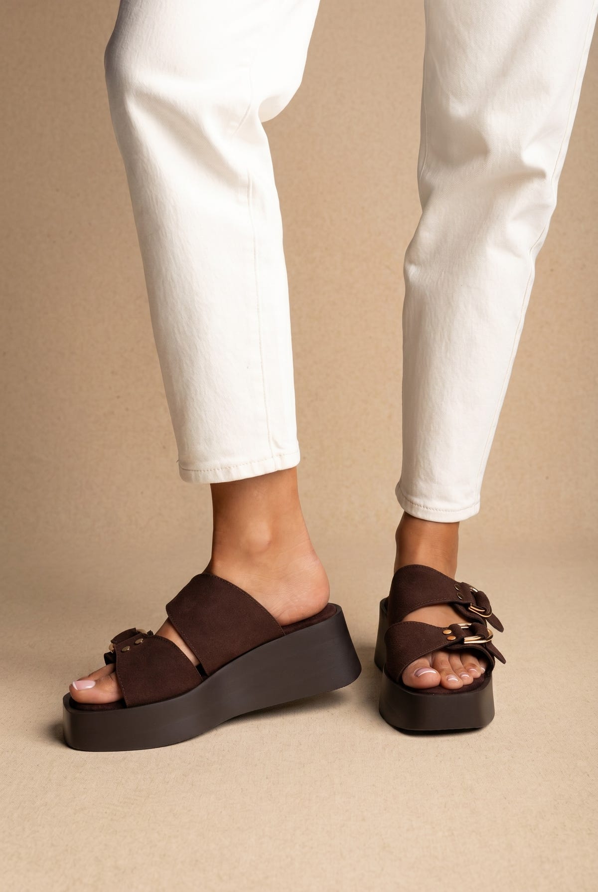 Dark brown suede wedge platform slides with double buckle straps, worn on a model with white jeans, front view