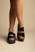 Black suede platform slides with two straps and gold ring buckles, worn on a model, front view of both feet