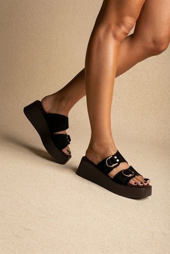 Black suede platform slides with gold ring buckles and dark brown platform soles, worn on a model, side view