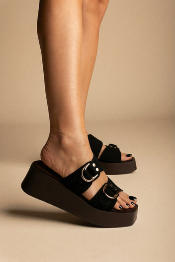 Black suede platform slides with two buckled straps and gold rings, worn on a model, side view against beige