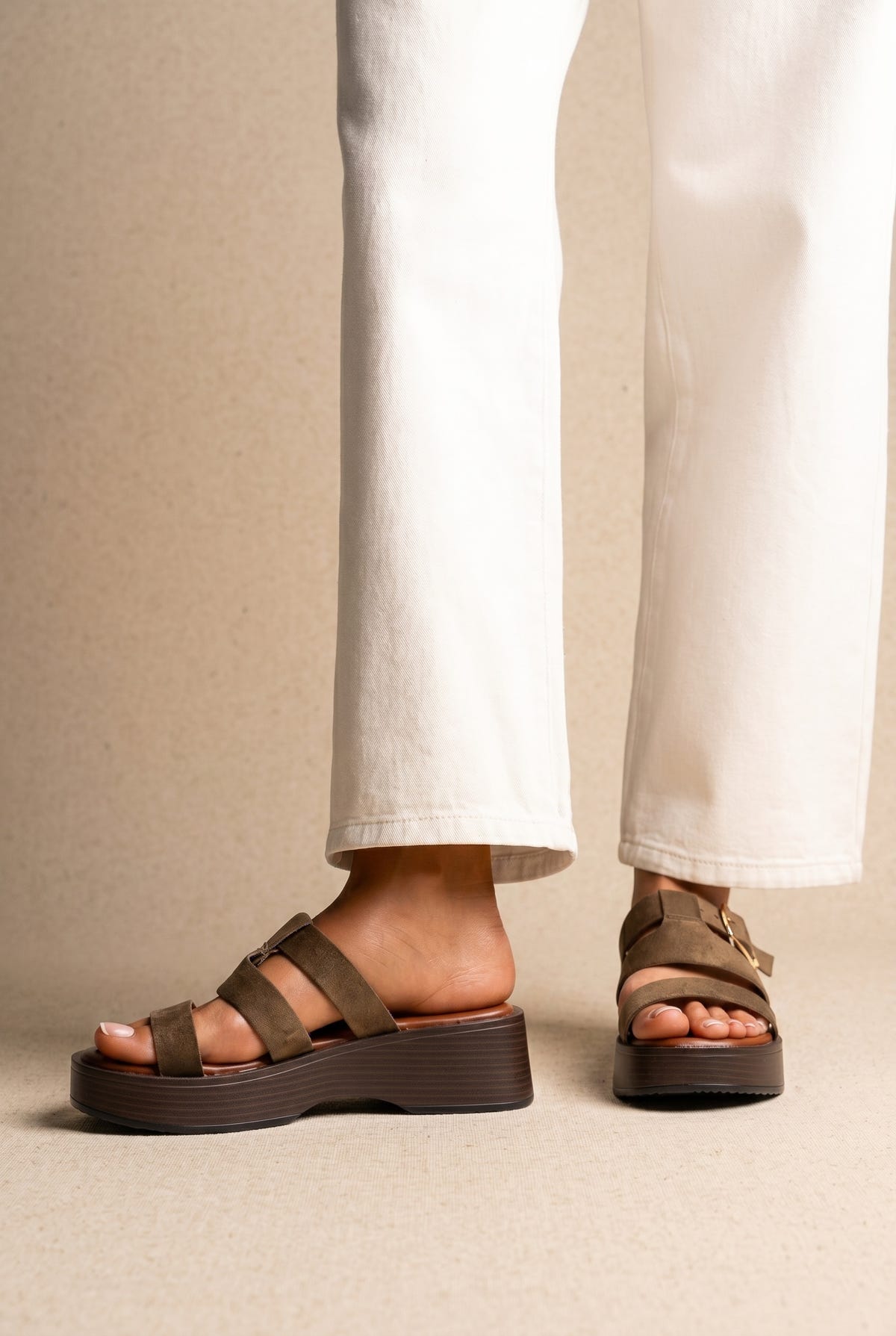 Olive suede platform slide sandals with wide straps and a chunky dark sole, worn on a model with white jeans, front view