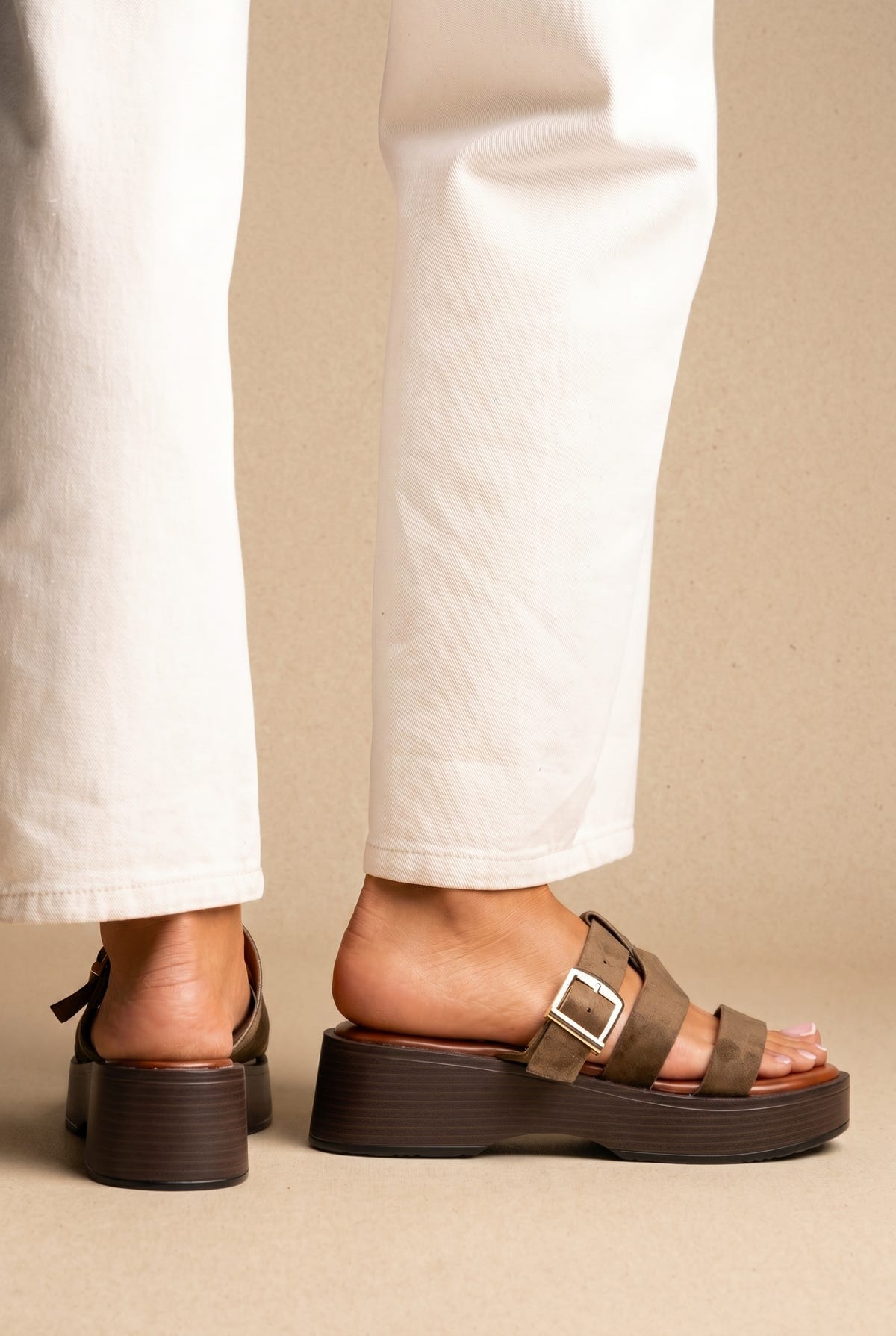 Olive suede platform slide sandals with a buckle strap and chunky dark brown sole, worn on a model with white jeans, side view