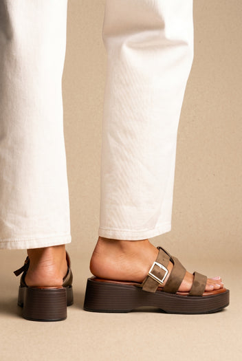 Olive suede platform slide sandals with a buckle strap and chunky dark brown sole, worn on a model with white jeans, side view
