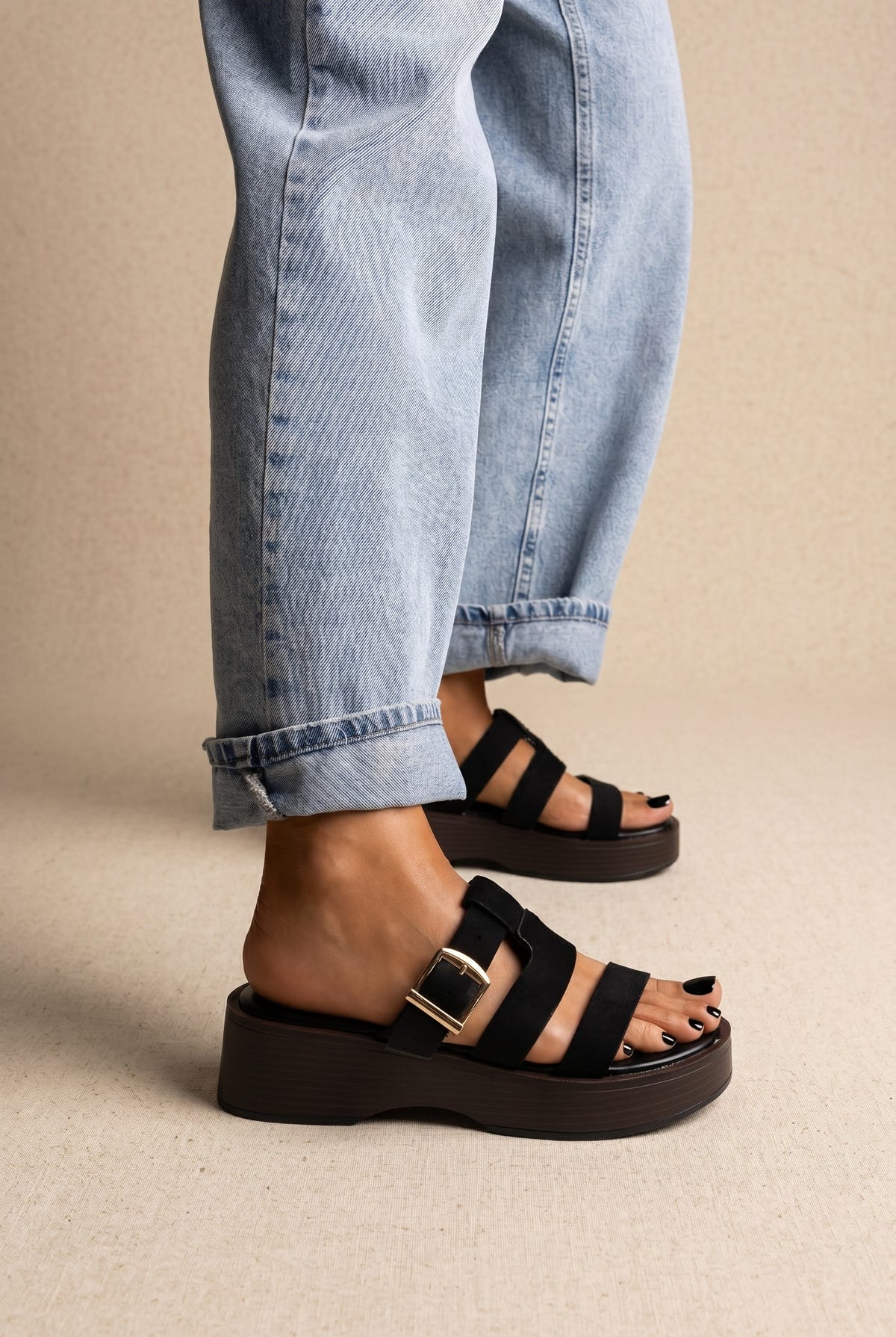Black suede platform slides with three straps and gold buckle on a dark brown sole, side view on a model