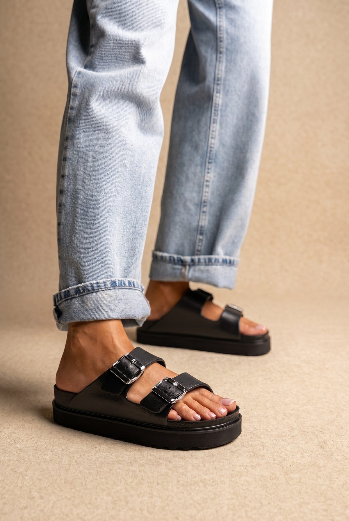 Black leather platform slide sandals with two silver buckle straps, worn on a model with light wash jeans, three-quarter view