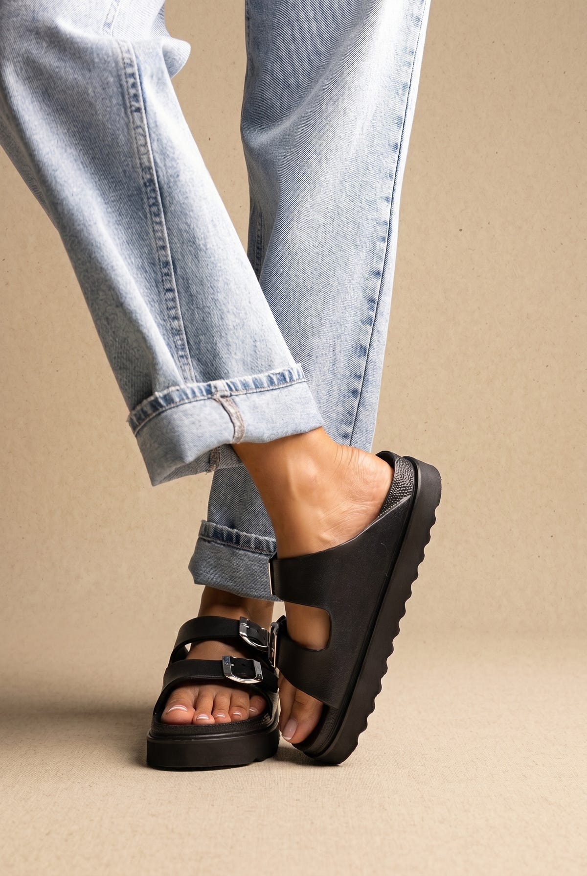 Black leather chunky platform slides with two buckle straps and a ridged sole, worn on a model with rolled jeans, side view