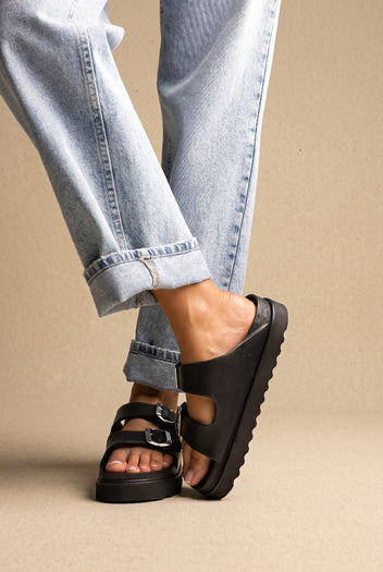 Black leather chunky platform slides with two buckle straps and a ridged sole, worn on a model with rolled jeans, side view