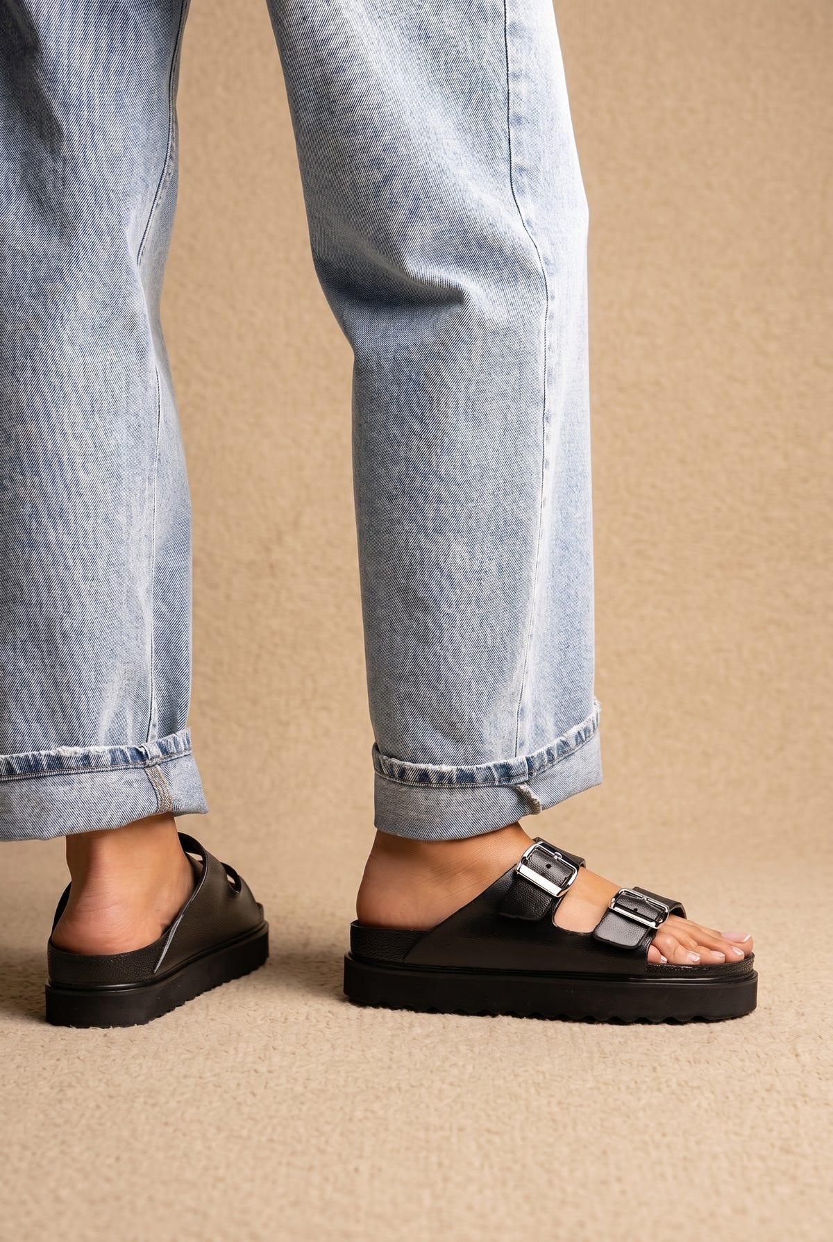 Black leather chunky platform slide sandals with double buckle straps, worn on a model with rolled light wash jeans, side view