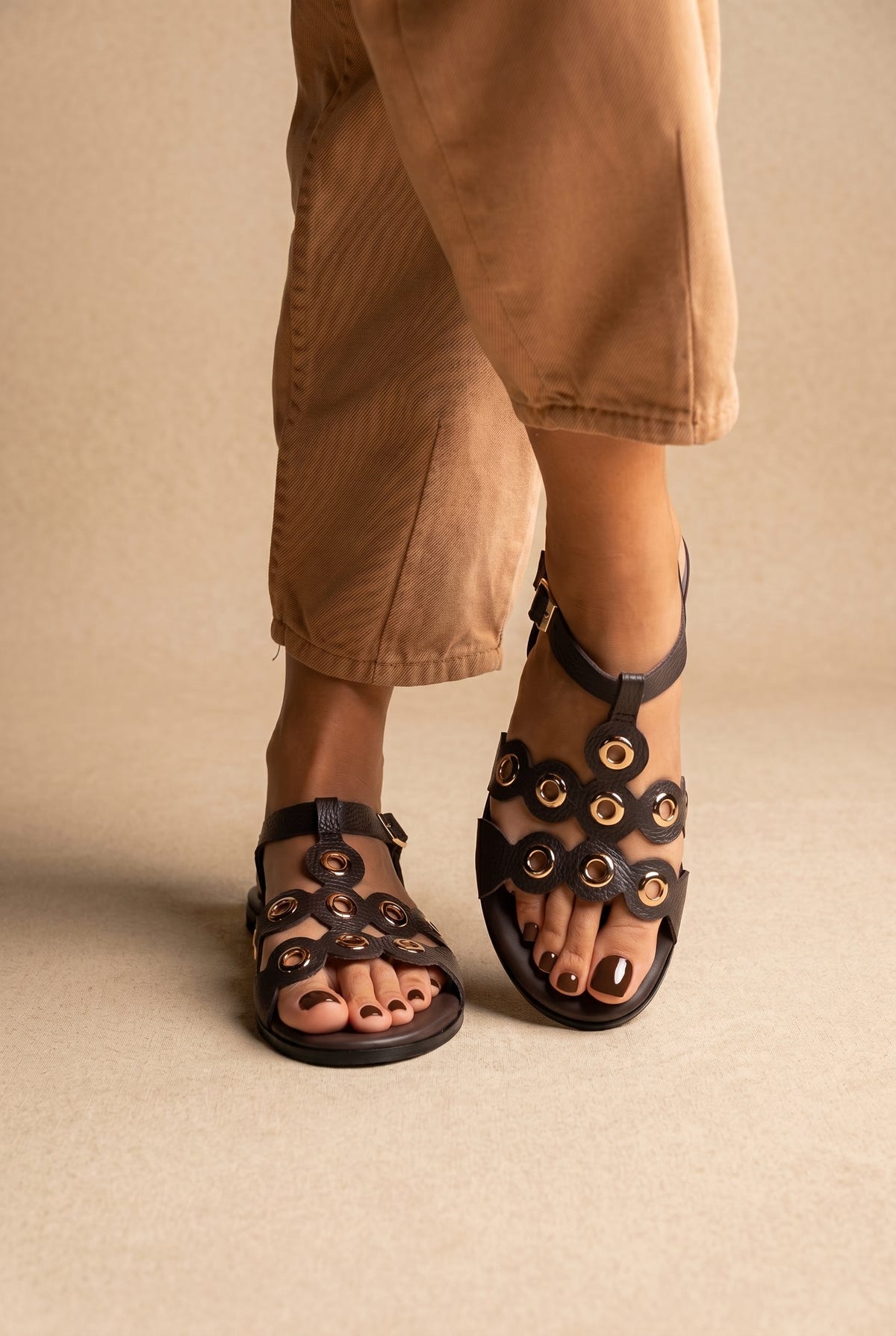 Dark brown leather flat sandals with gold eyelet circles and ankle strap, front view worn with tan trousers