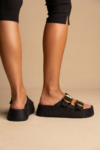 Black woven raffia platform slides with two gold buckles, worn on a model in dark trousers, side and back view