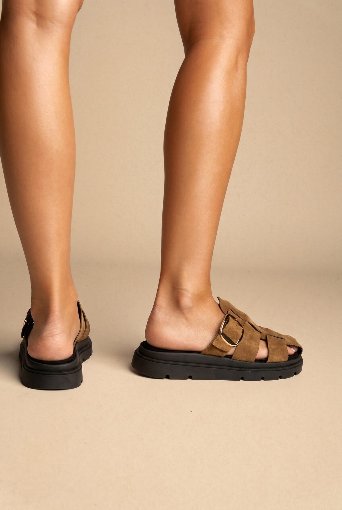 Tan suede woven strap slide sandals with gold buckle and chunky black sole, worn on a model, back view