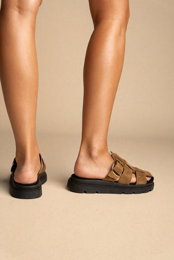 Tan suede woven strap slide sandals with gold buckle and chunky black sole, worn on a model, back view