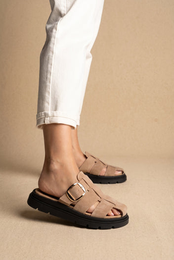 Beige suede caged slide sandals with gold buckle and thick black sole, worn with white jeans, side view