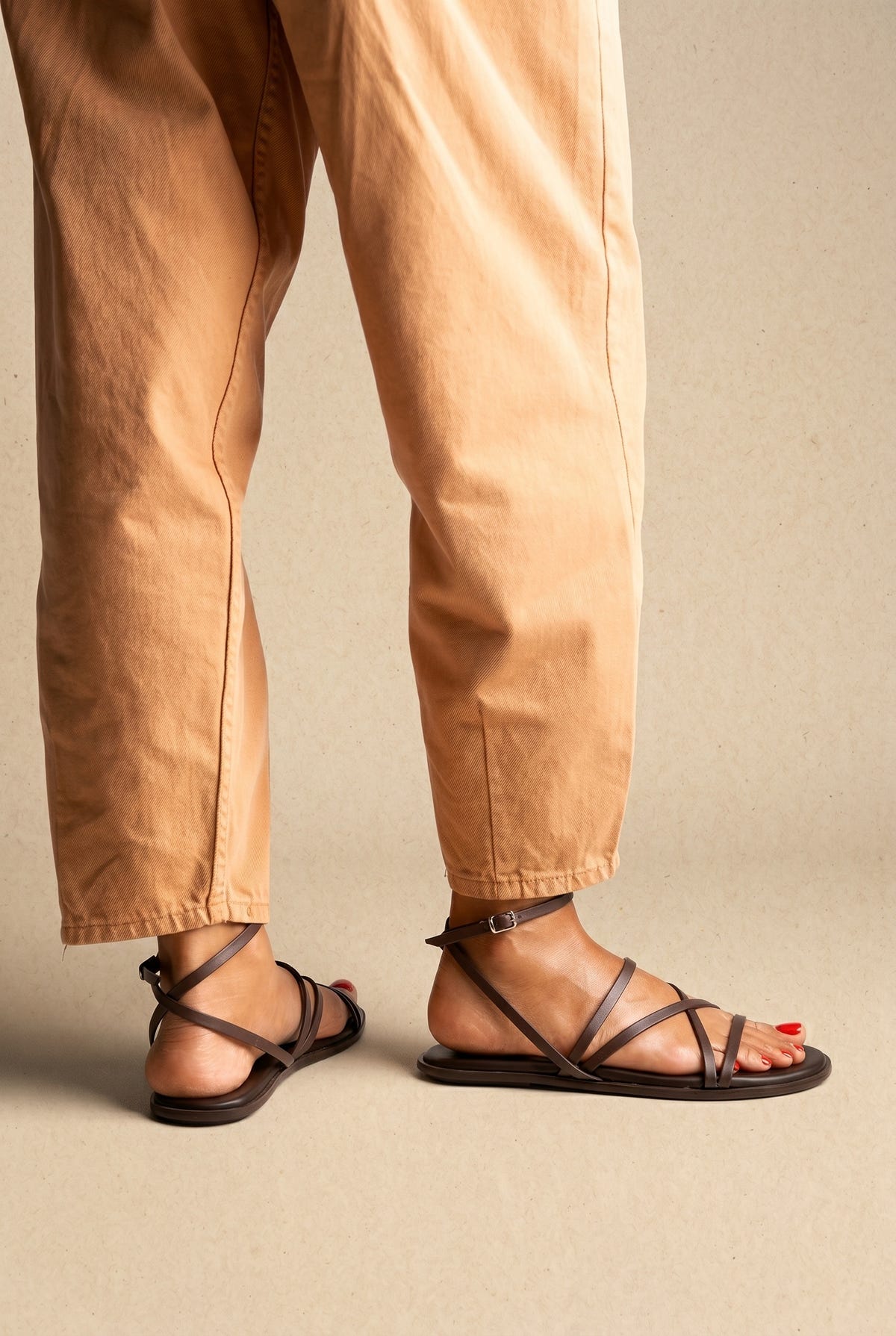Dark brown leather flat strappy sandals with thin crossing straps and ankle buckle, back and side view on a model