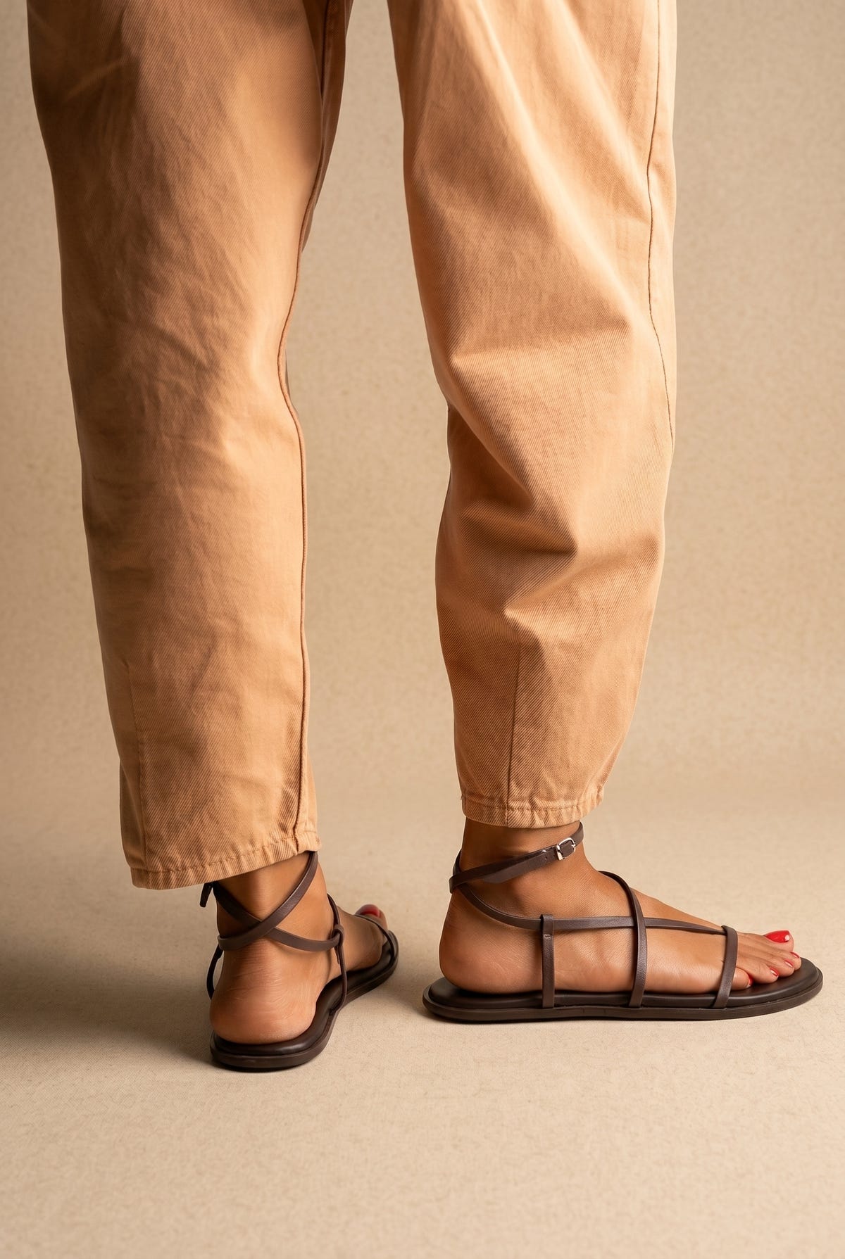 Dark brown leather flat strappy sandals with parallel thin straps and buckled ankle strap, back view on a model