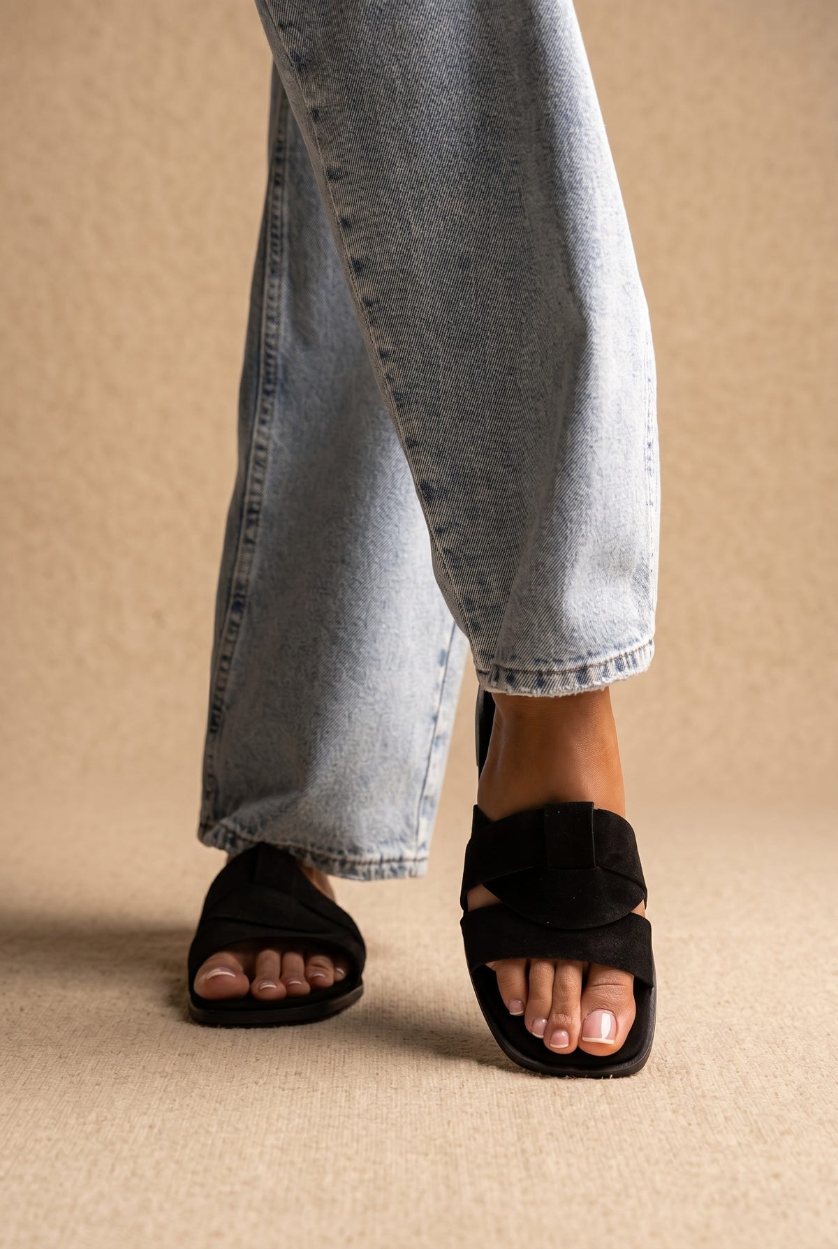 Black suede flat slide sandals with wide woven crossover straps, worn on a model with light wash wide-leg jeans, front view
