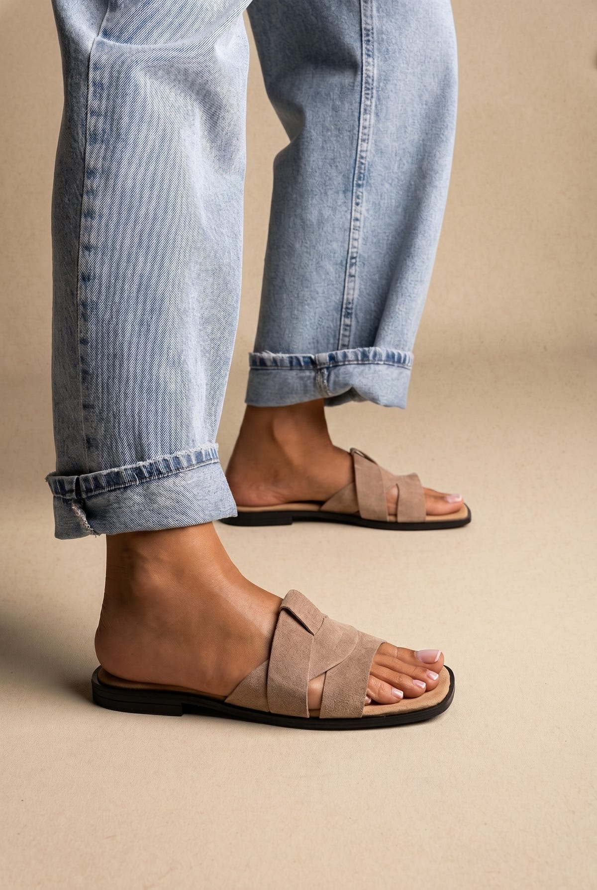 Taupe suede flat slides with twisted crossover straps and black sole, side view on a model in rolled jeans