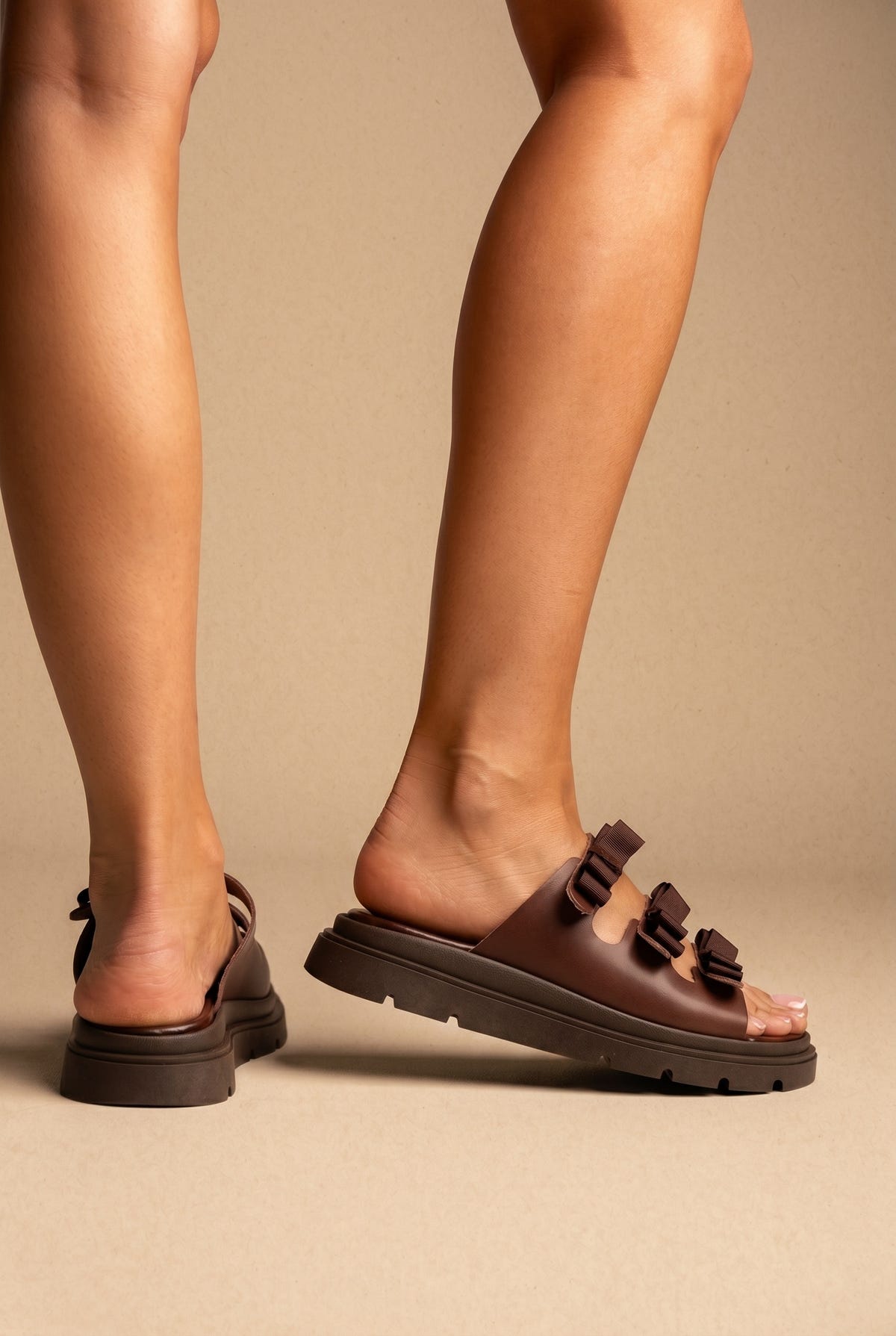 Brown leather chunky platform slides with three bow-style straps on a thick dark brown lugged sole, worn on a model