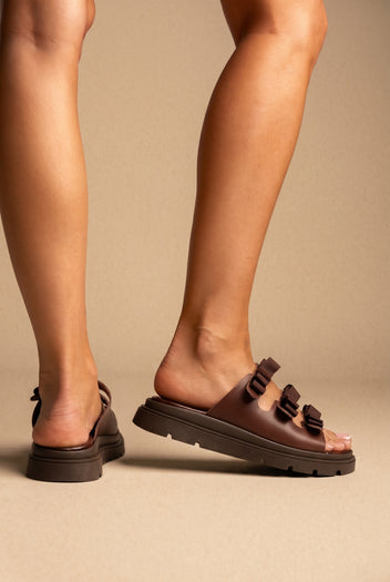 Brown leather chunky platform slides with three bow-style straps on a thick dark brown lugged sole, worn on a model
