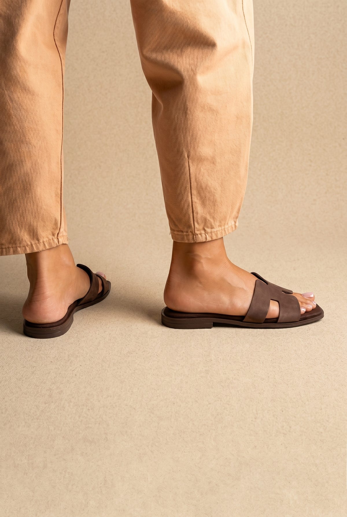 Dark brown flat H band slides with brown flat sole, back and side view on a model in tan trousers
