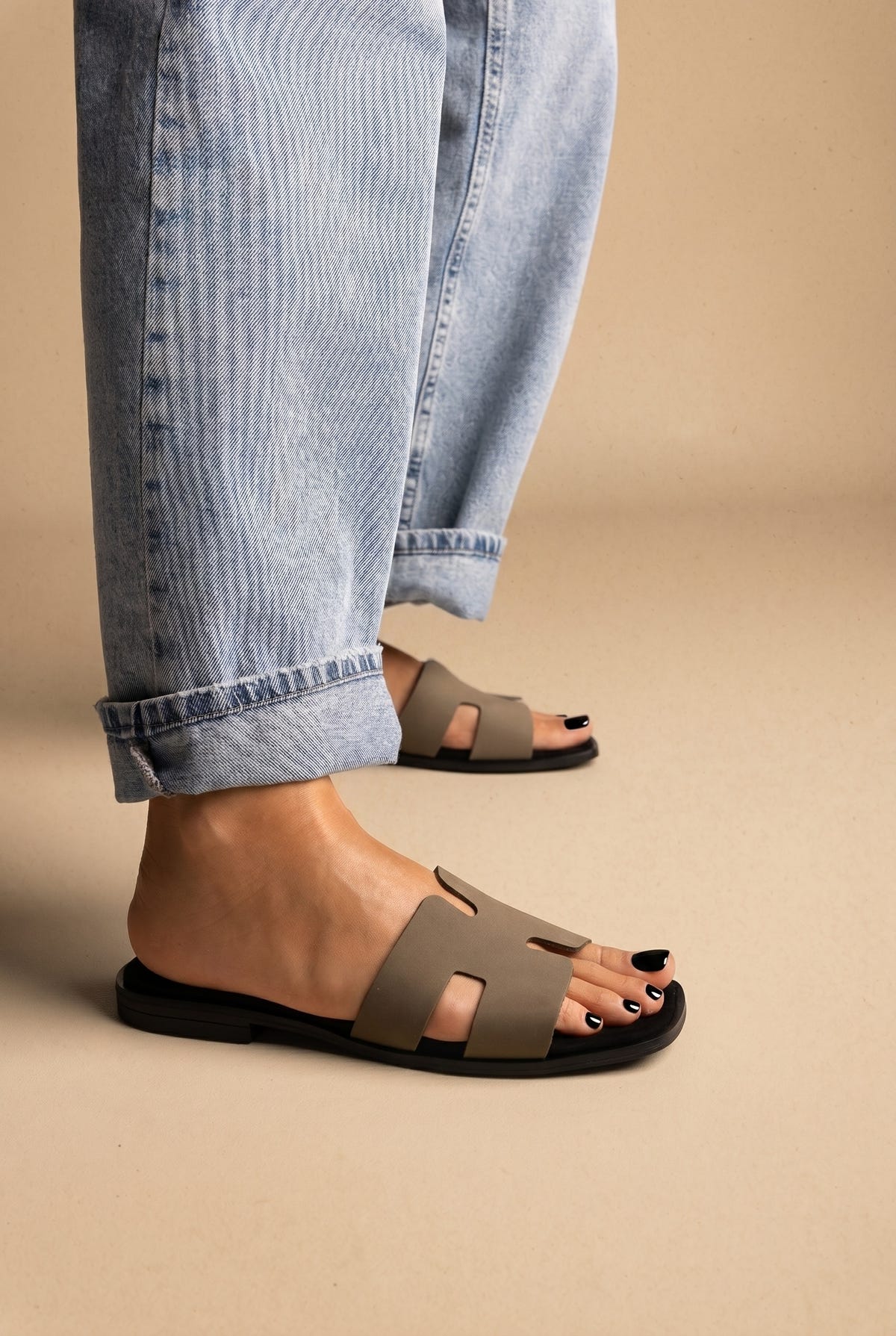 Taupe grey H band flat slides with black sole, side view on a model in rolled light blue jeans