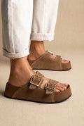 Taupe suede platform slides with two gold buckles and cream stitching, side view of a model wearing both shoes