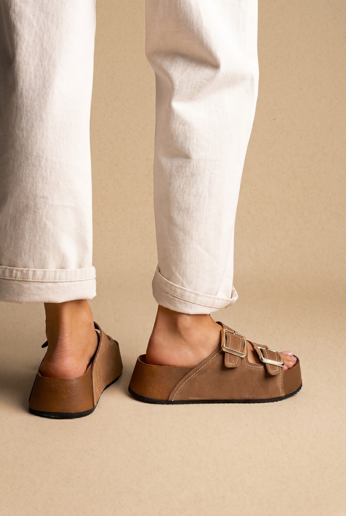 Taupe suede platform slides with two gold buckle straps, side and back view worn on a model in white cuffed trousers