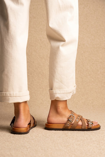 Tan suede flat sandals with multiple buckled straps and studs on a cork-style footbed, worn on a model with cream jeans