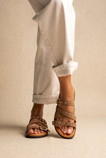 Tan suede studded buckle strap flat sandals on a tan footbed with dark sole, worn on a model with cuffed cream jeans