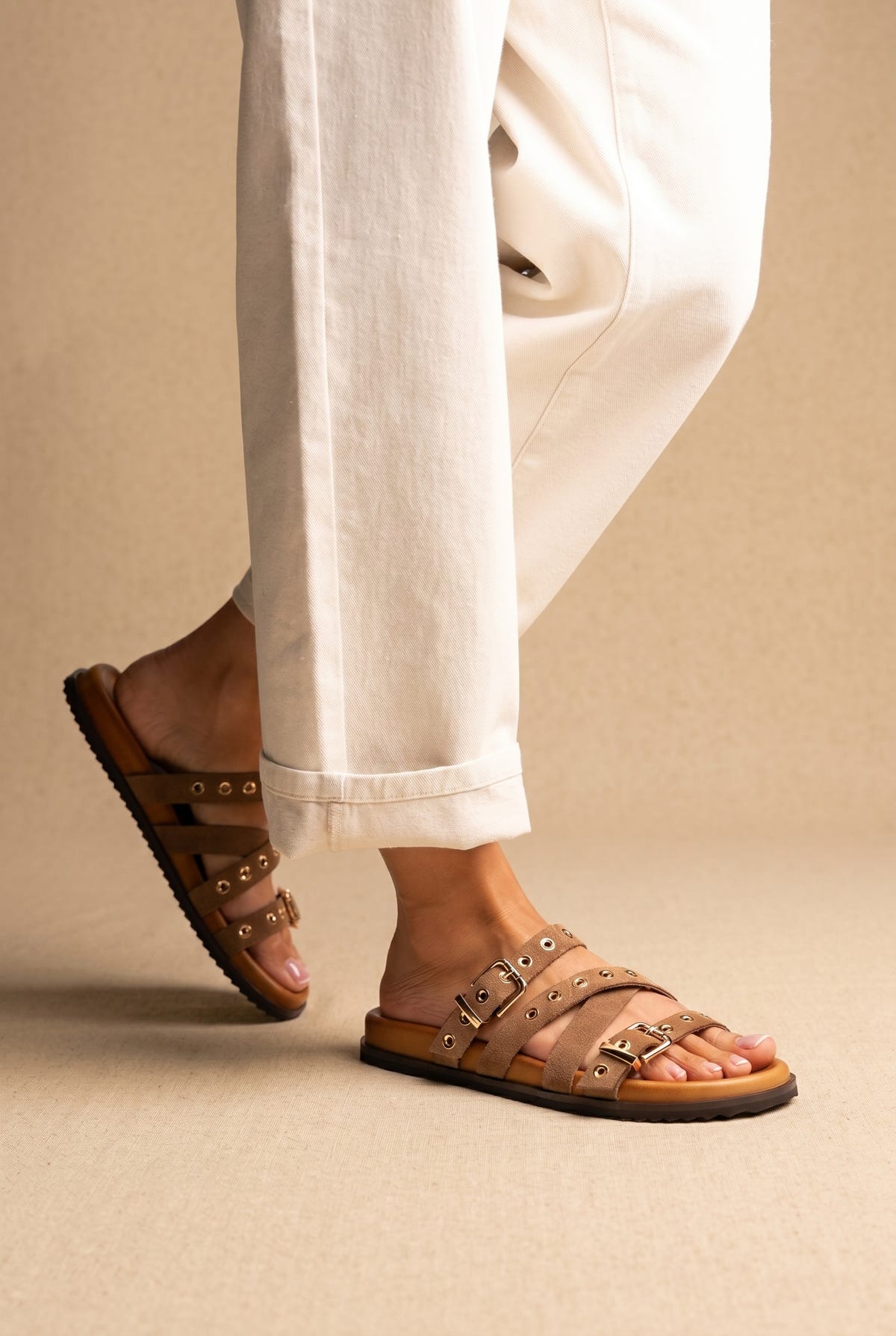 Tan suede flat sandals with buckled eyelet straps and tan footbed, worn on a model with cream trousers, side view