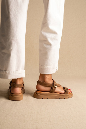 Tan suede chunky platform sandals with gold buckles and a thick lug sole, worn on a model with cream trousers, back view