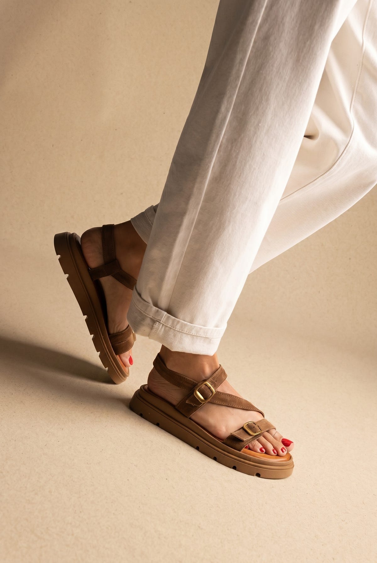 Tan suede chunky sandals with gold buckle straps and lug sole, one foot raised, worn on a model with cream trousers