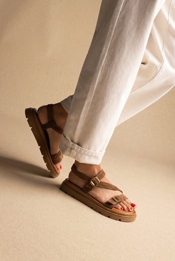 Tan suede chunky sandals with gold buckle straps and lug sole, one foot raised, worn on a model with cream trousers