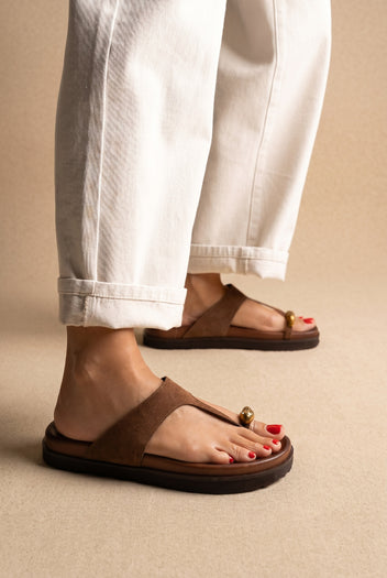 Brown suede toe-post slide sandals with gold toe ornament and thick sole, worn on a model with white cuffed trousers