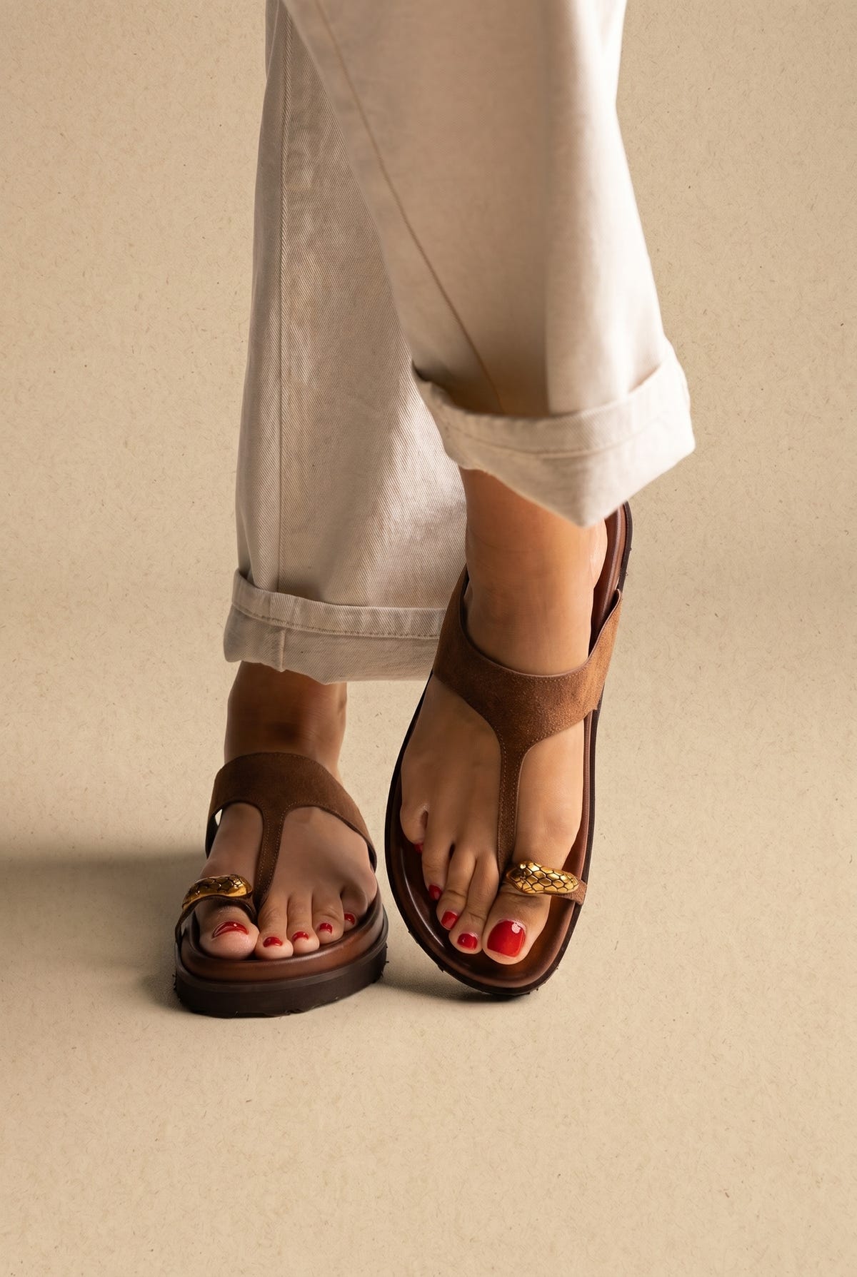 Brown suede thong sandals with gold toe ring detail and chunky sole, front view on a model with white trousers