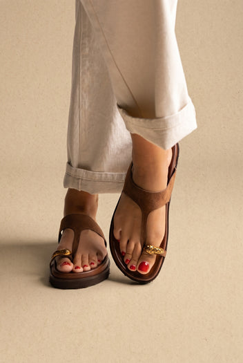 Brown suede thong sandals with gold toe ring detail and chunky sole, front view on a model with white trousers
