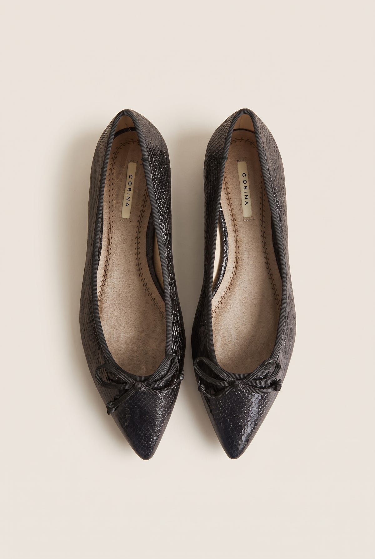 Top-down view of pair of black snake-embossed pointed flats with small bows and tan lining