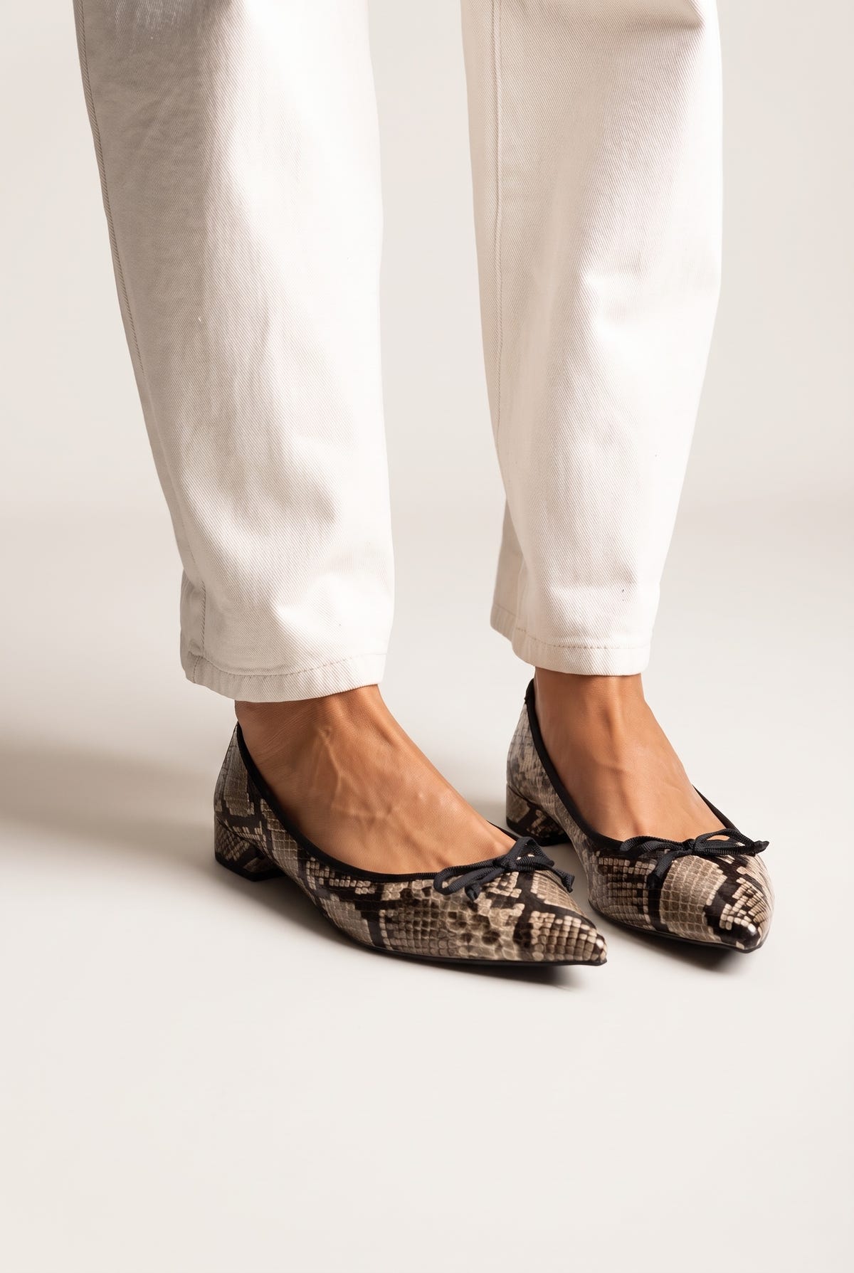Snake-embossed leather bow-detail ballet flats, pair in front view on a model wearing white trousers in studio light