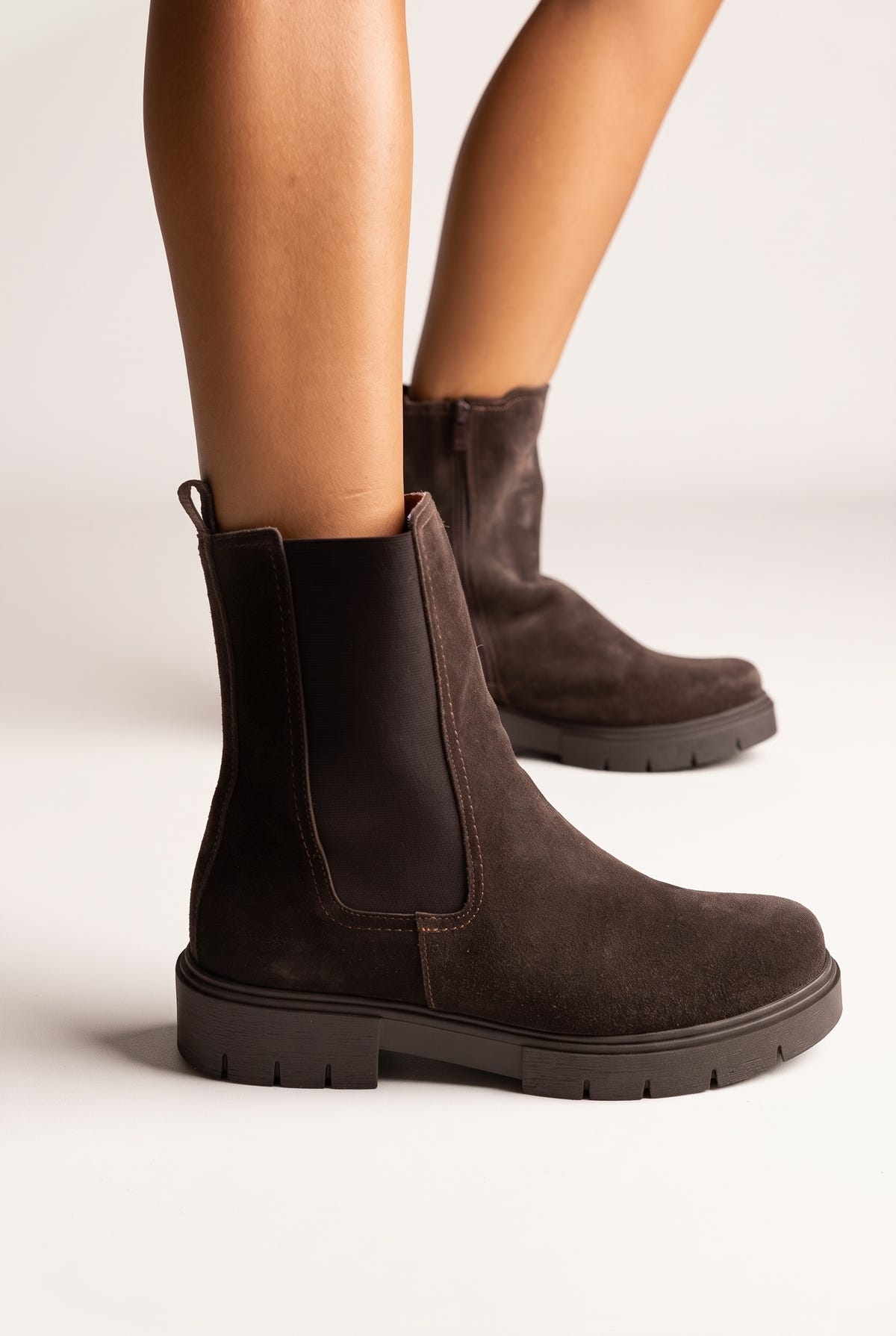Model legs in dark brown suede chelsea ankle boots with elastic side panels, angled view on light beige background
