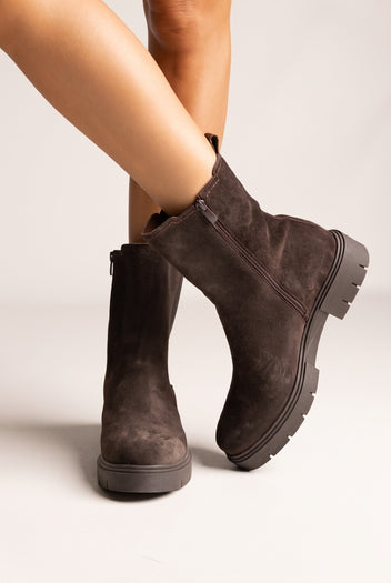 Model legs in dark brown suede chelsea ankle boots with lug sole, angled view on light beige background