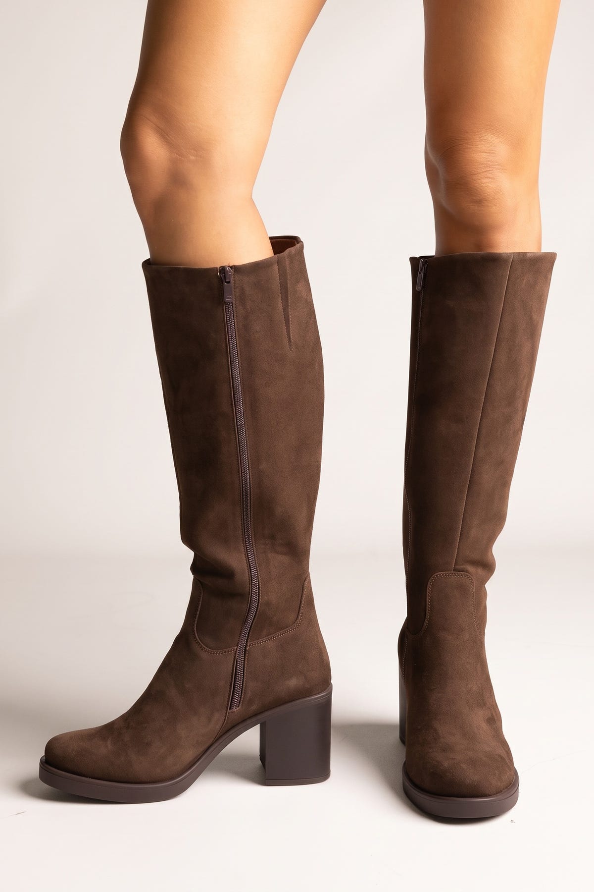 Pair of brown suede knee-high block heel boots with smooth shafts and block heels, front view on a light beige background
