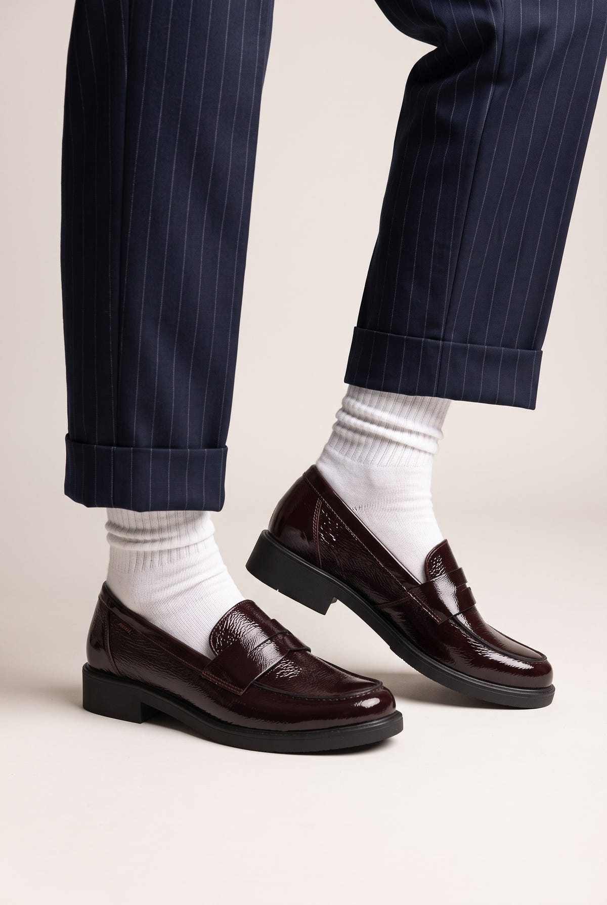 Model legs in burgundy patent penny loafers with white socks and navy trousers, angled view on light grey