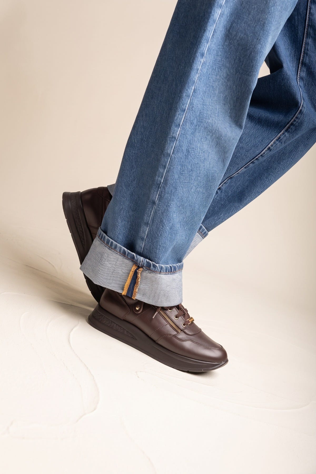Model in rolled up jeans wearing brown leather wedge sneakers with laces and a chunky sole turned to show the toe