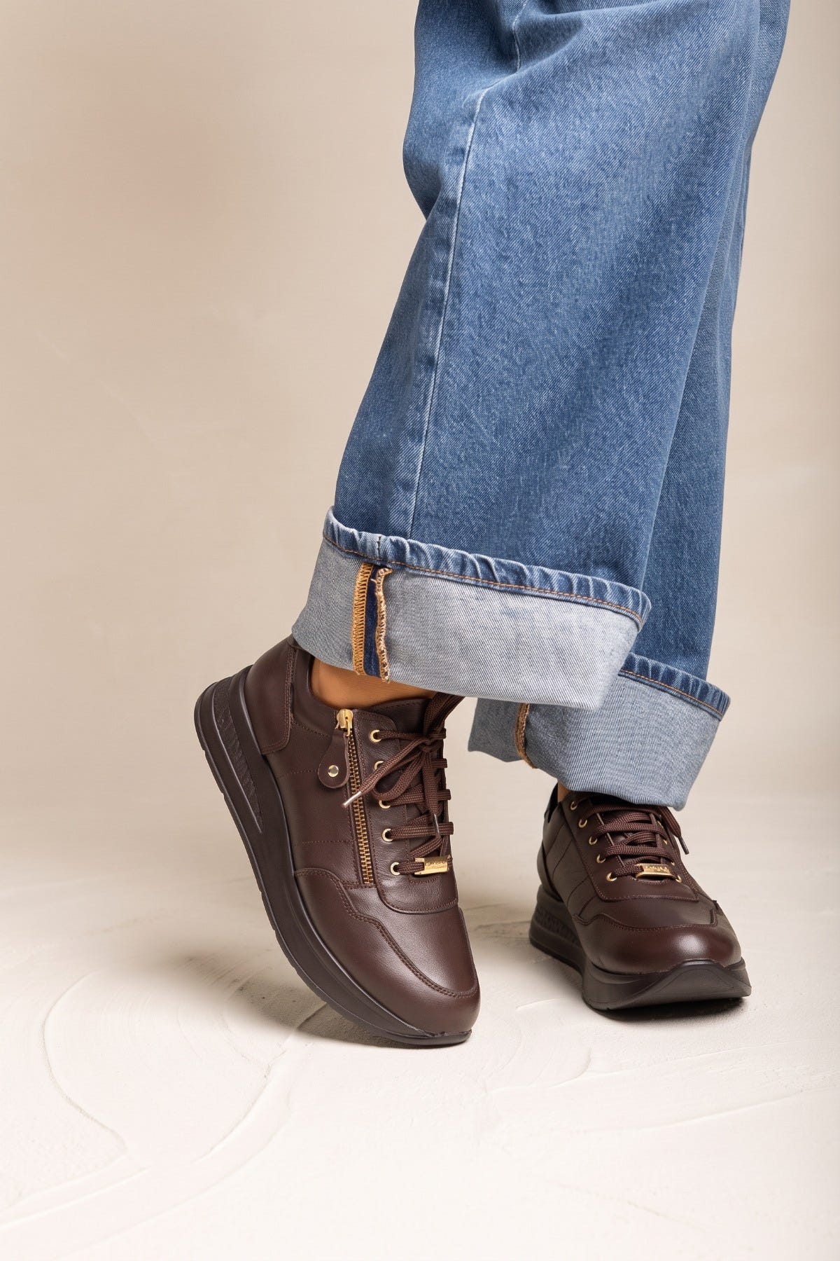Model wearing brown leather wedge sneakers from a three quarter front angle showing the laces and gold tone side detail